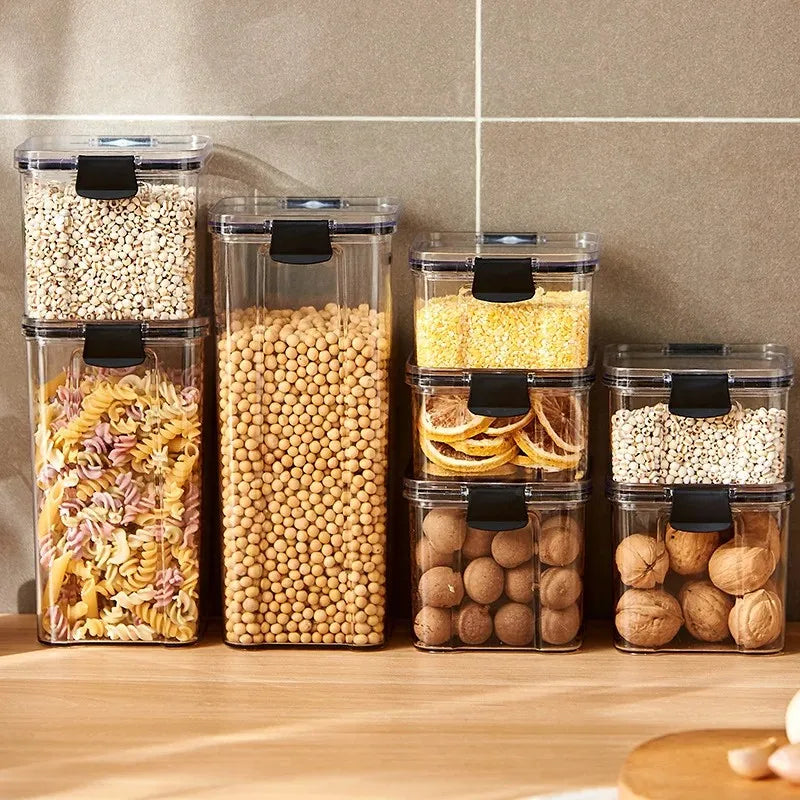 A collection of transparent storage containers filled with various dry foods like pasta, beans, nuts, and cereals on a wooden surface.