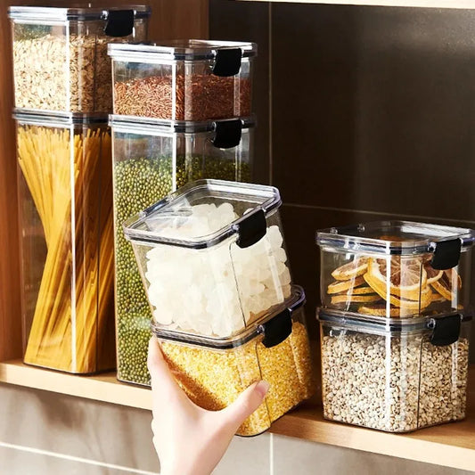 A collection of transparent storage containers filled with various dry foods like pasta, beans, nuts, and cereals on a wooden surface.