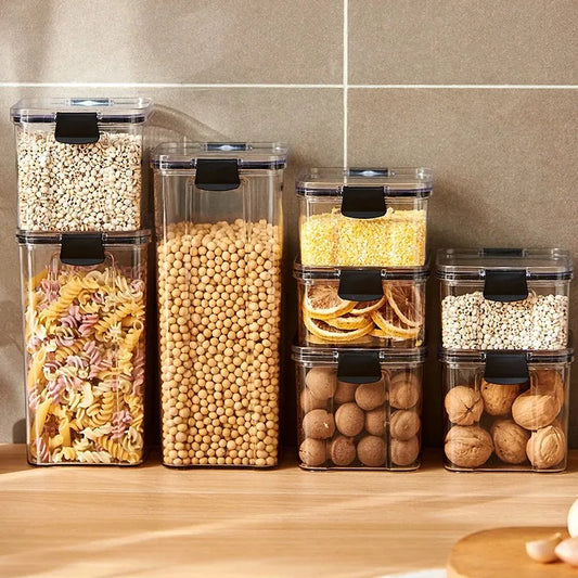 A collection of transparent storage containers filled with various dry foods like pasta, beans, nuts, and cereals on a wooden surface.